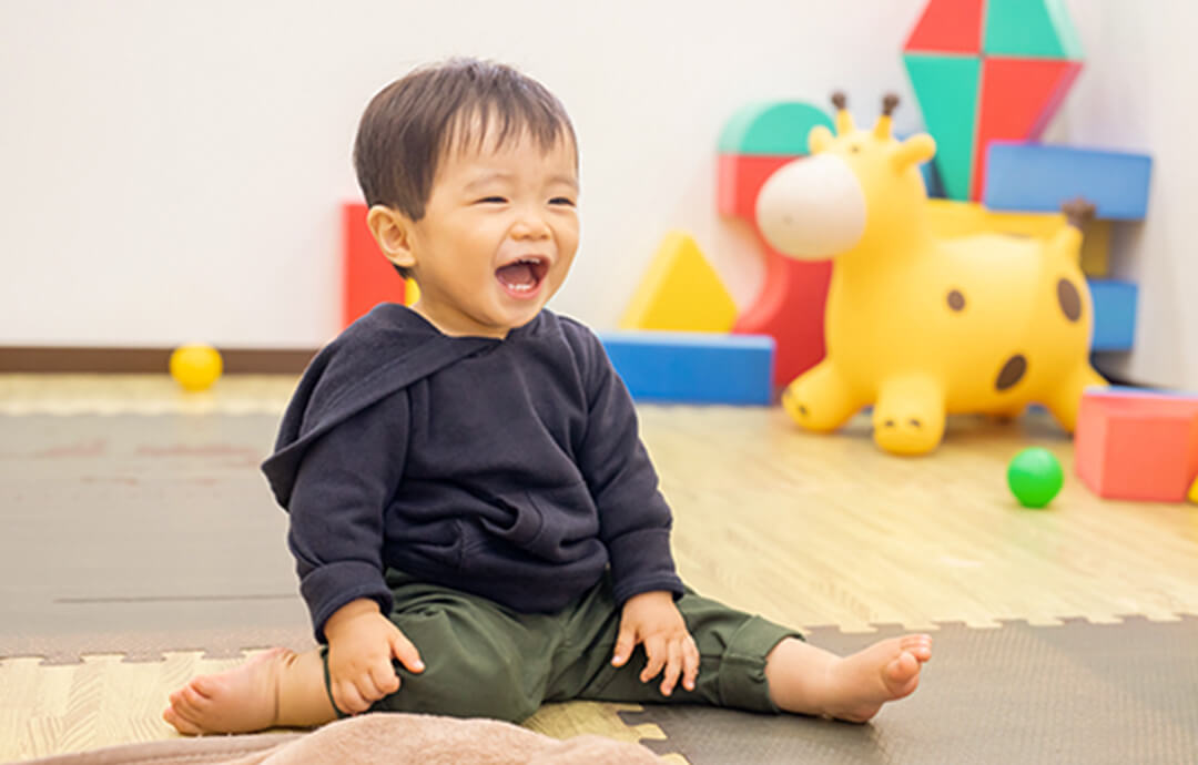 室内のプレイマットの上で、満面の笑みで遊んでいる幼い男の子の写真。
