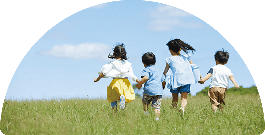 屋外の広々とした緑の野原で子どもたちが4人、手をつないで晴れた空に向かって楽しそうに走り去っている後ろ姿。
